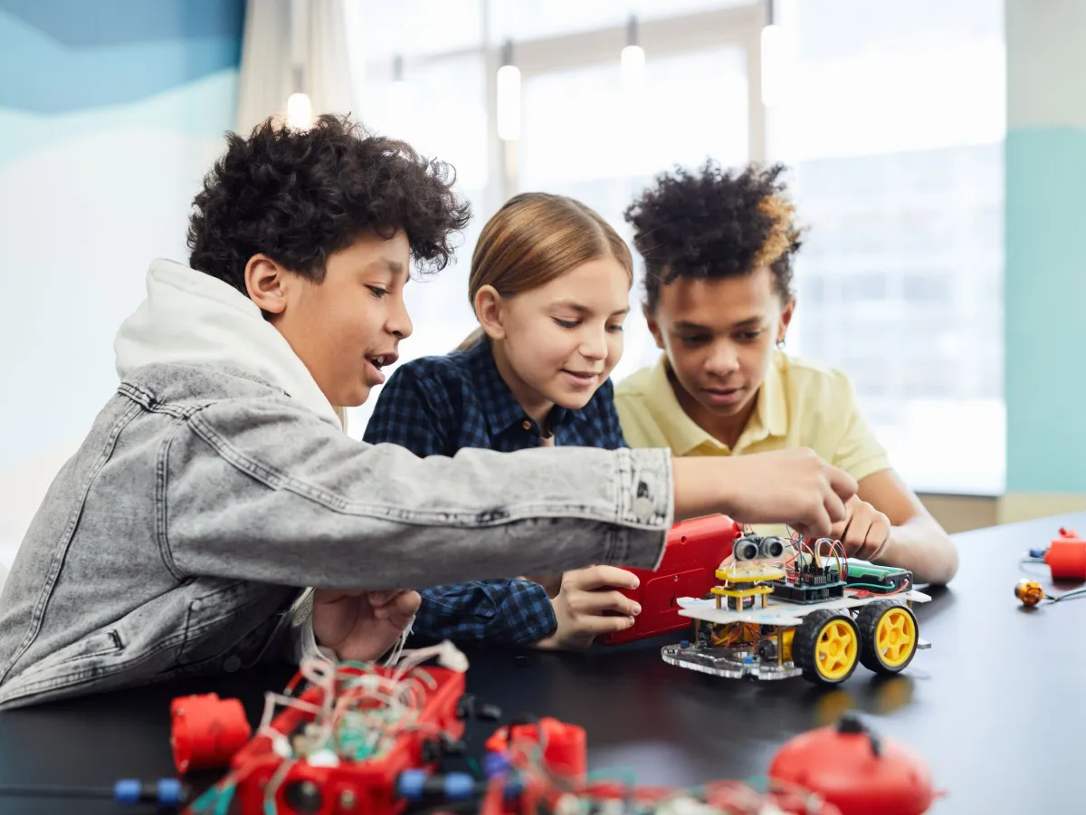Three kids building and wiring a robot car together in a bright robotics classroom