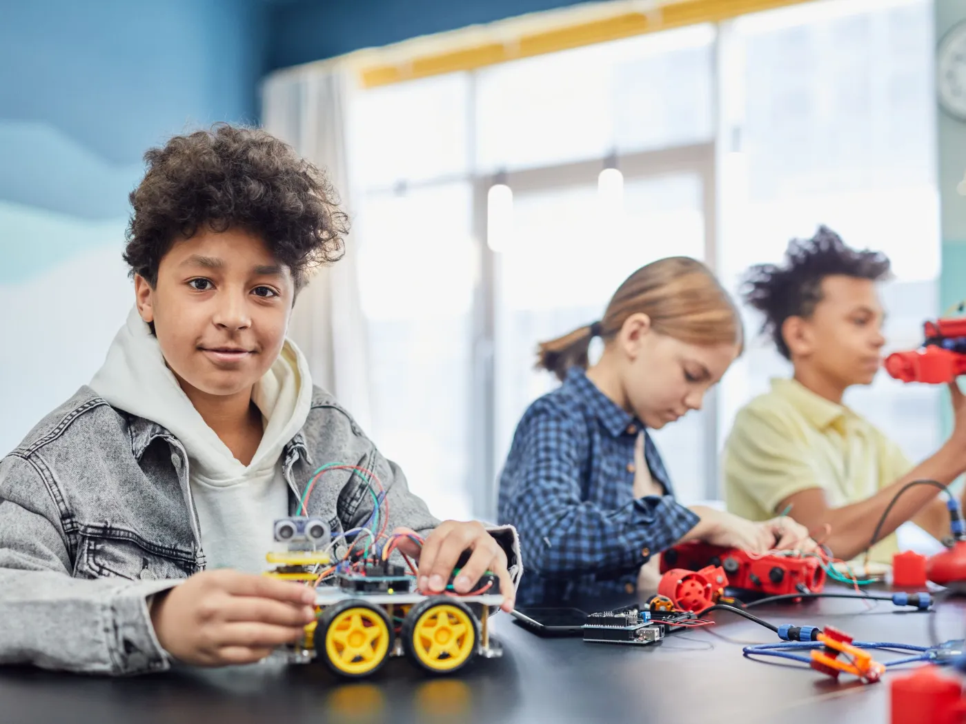 Smiling boy with the robot car he built, classmates working on robotics kits behind him